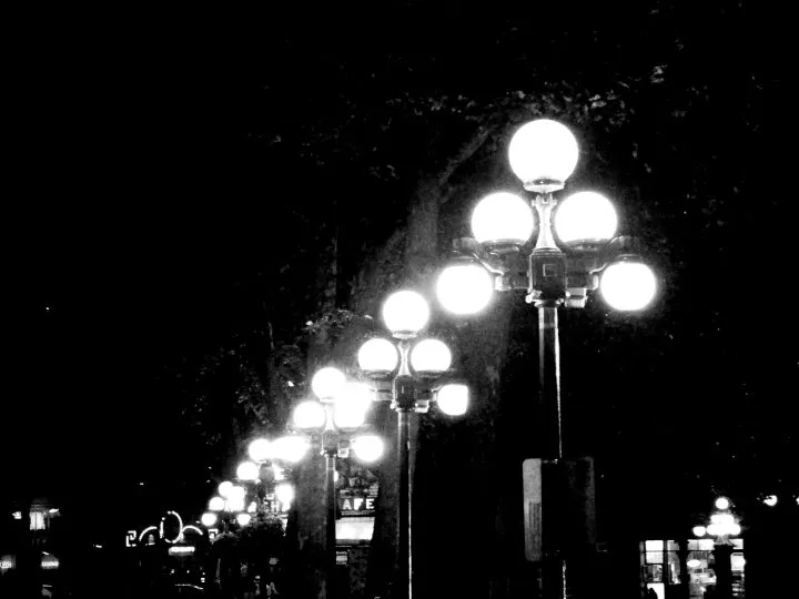 A receding row of five-globe cast-iron street lamps glowing at night in Pioneer Square, shot in high-contrast black and white, with a lit cafe sign further down the block