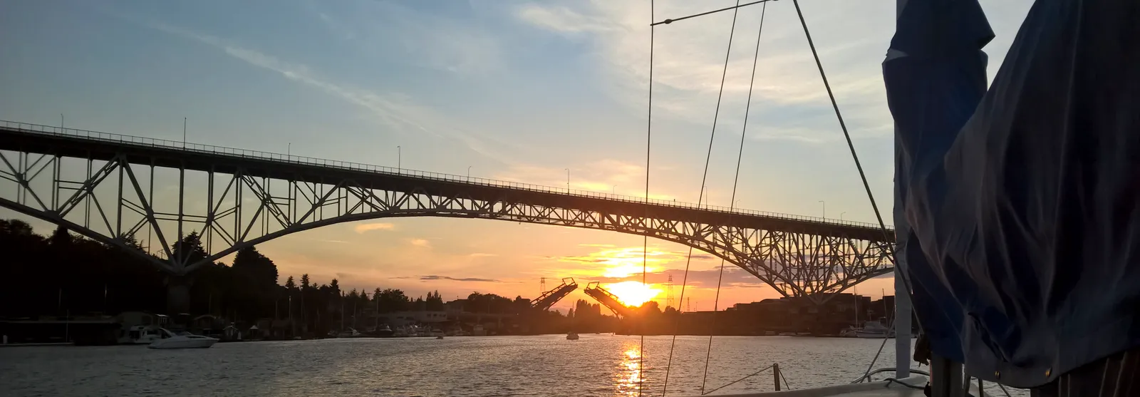 Looking west along the ship canal from a sailboat at sunset, the steel arch of the Aurora Bridge overhead, the Fremont Bridge raised with both leaves up and the sun setting through the gap, rigging and a furled sail in the right foreground