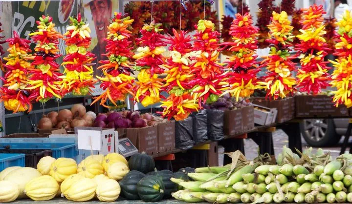 A Pike Place Market produce stall, chile ristras hanging in red, orange and yellow above crates of delicata and acorn squash, sweet corn and onions