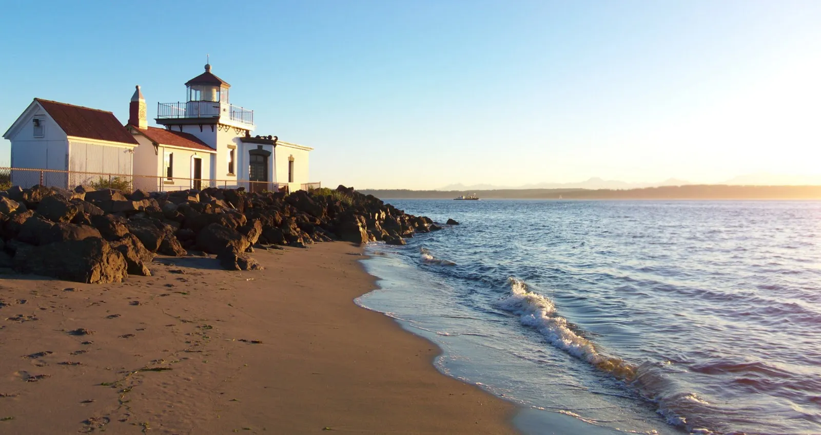 West Point Lighthouse at Discovery Park in low golden light, white walls and a red roof above a riprap seawall, with sand beach and small surf in the foreground and the Olympic peninsula faint across the water
