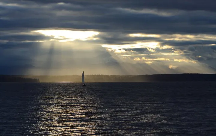 A single sailboat on Puget Sound off Shilshole Bay, lit from behind as the sun breaks through a heavy cloud deck in visible shafts, with the far shore low on the horizon