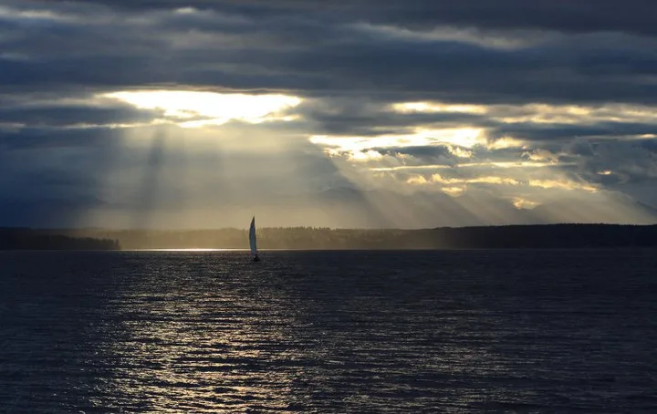 A single sailboat on Puget Sound off Shilshole Bay, lit from behind as the sun breaks through a heavy cloud deck in visible shafts, with the far shore low on the horizon