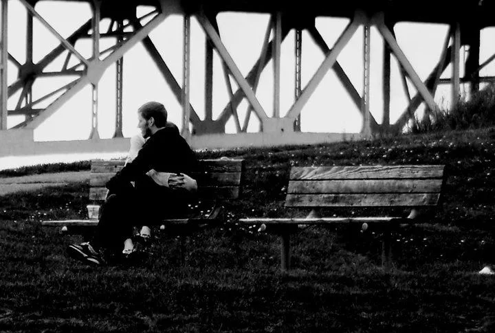 A couple embracing on a park bench beneath a steel truss bridge, an empty bench beside them