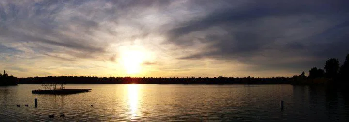 A wide view of Green Lake at sunset in late October, a heavy cloud deck with the sun burning through low, a swimming float on the left and ducks on the water