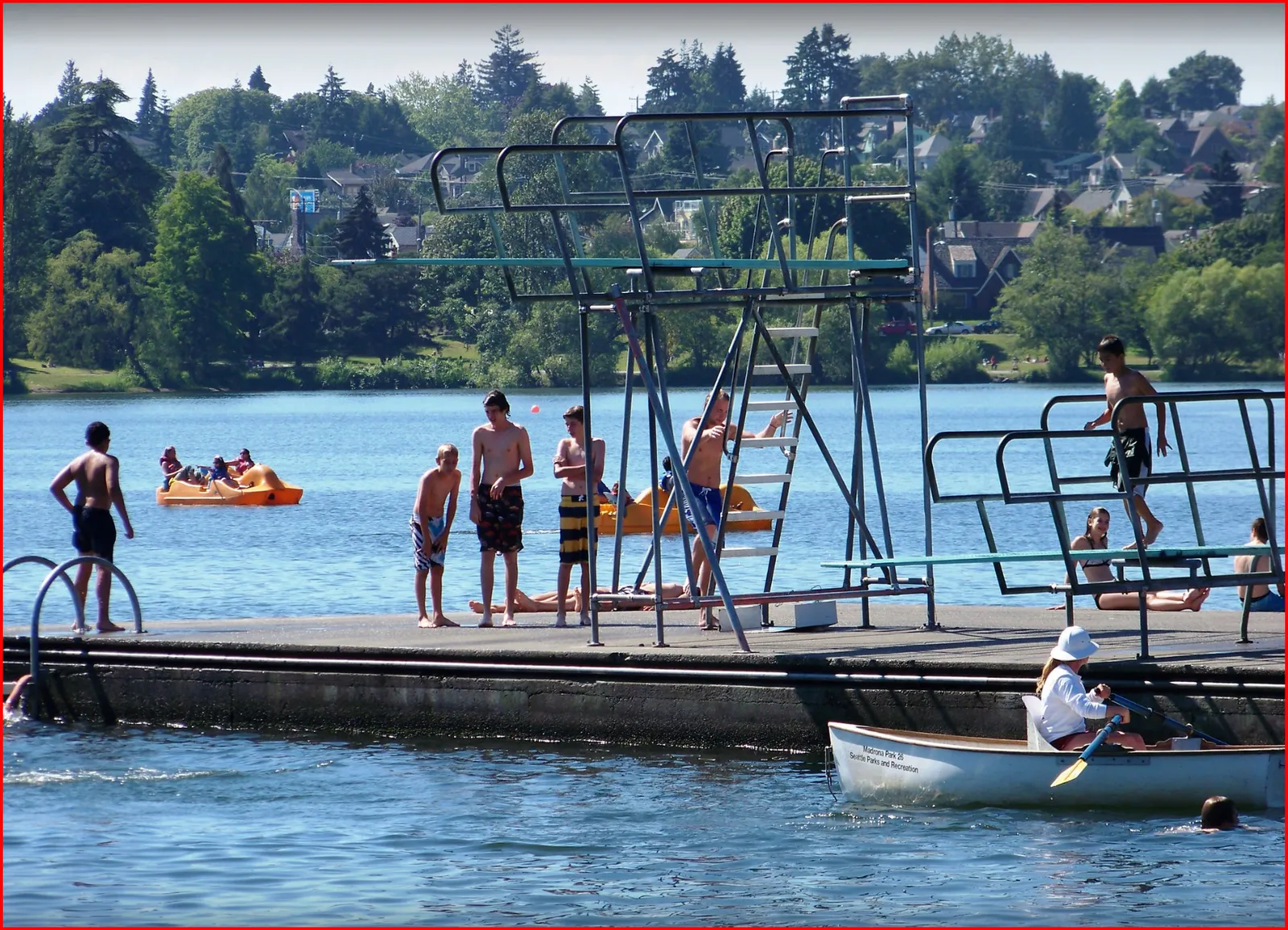 The swimming raft at Green Lake in summer, teenagers standing along the deck waiting their turn at the diving boards, pedal boats on the water behind and a lifeguard rowboat in the foreground