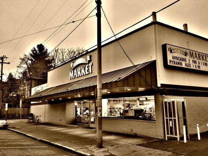 The Montlake Boulevard Market at dusk in warm sepia tone, a low corner store with a lit interior, a reader board advertising avocados two for one dollar and Pyramid Ales at six ninety-nine, a bicycle parked outside